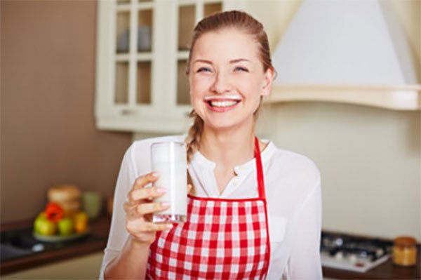 A woman in an apron with a glass of kefir in her hand and a trace of it above her upper lip